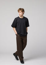 A young man with light brown hair wears IM MEN’s TURN: a loose black jersey T-shirt, wide brown trousers, and black shoes. Hands in pockets, he walks against a plain light gray background, embodying modern simplicity.