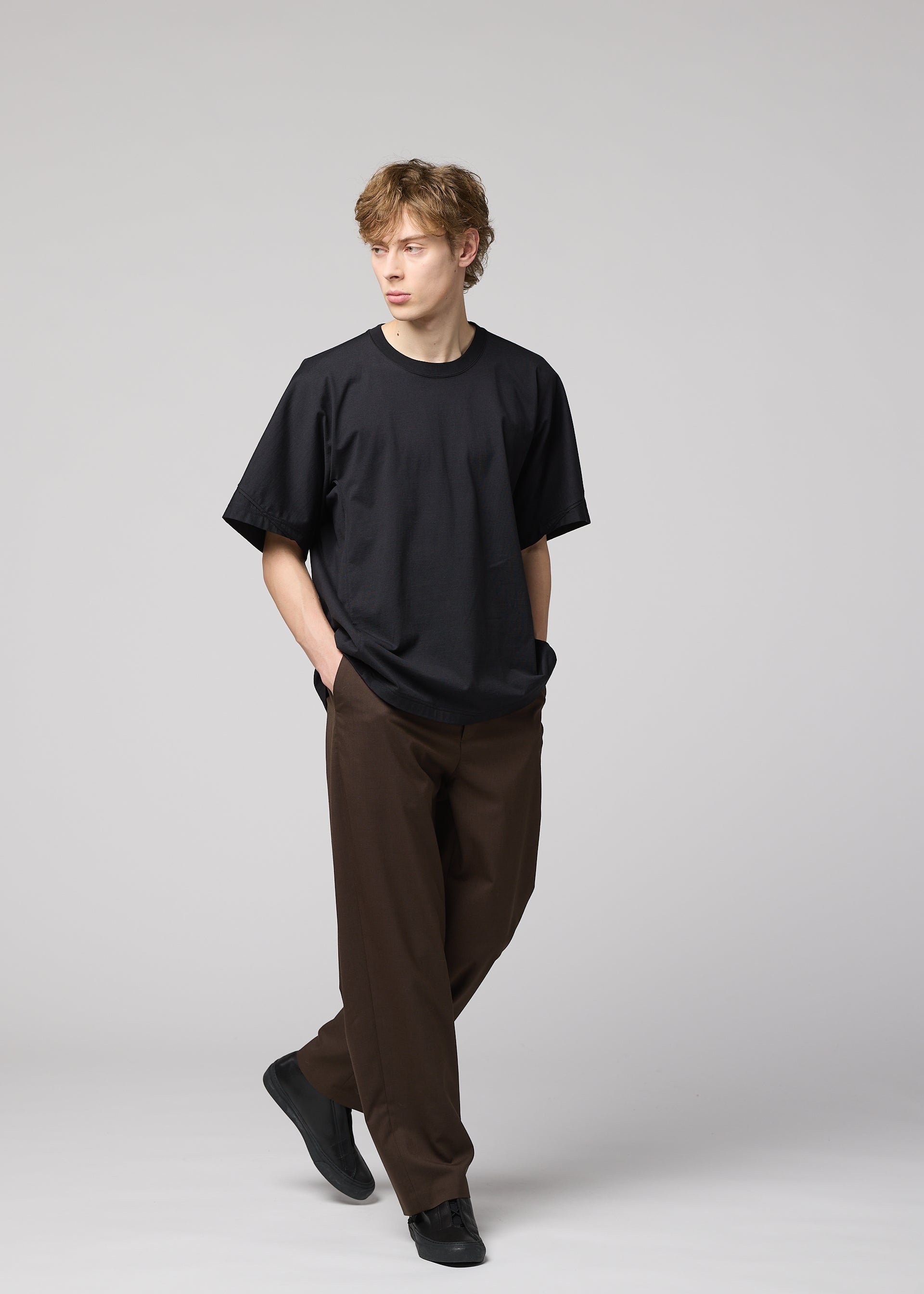 Zoomed view A young man with light brown hair wears IM MEN’s TURN: a loose black jersey T-shirt, wide brown trousers, and black shoes. Hands in pockets, he walks against a plain light gray background, embodying modern simplicity.