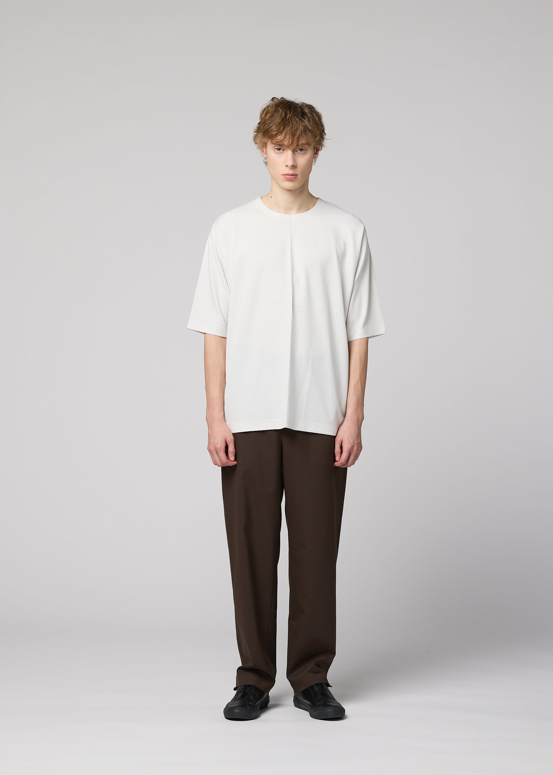 Zoomed view A person stands against a plain gray background, wearing the IM MEN FLAT T (an easy-care white short-sleeve jersey), dark brown crease-front pants, and black shoes, looking directly at the camera with a neutral expression.