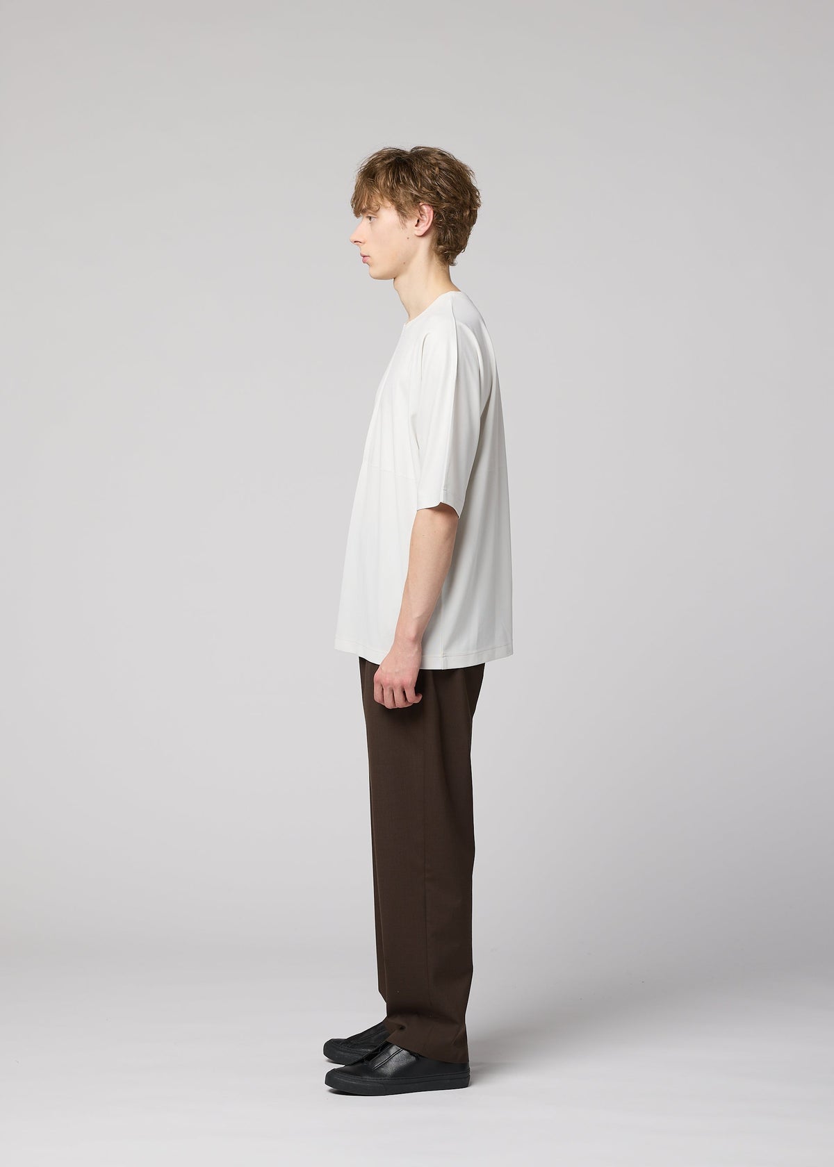 A young person with short light brown hair stands in profile against a plain light background, wearing the IM MEN FLAT T easy-care jersey pressed T-shirt, dark brown pants with bold creases, and black shoes. view 8 of 9