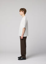 A young person with short light brown hair stands in profile against a plain light background, wearing the IM MEN FLAT T easy-care jersey pressed T-shirt, dark brown pants with bold creases, and black shoes.