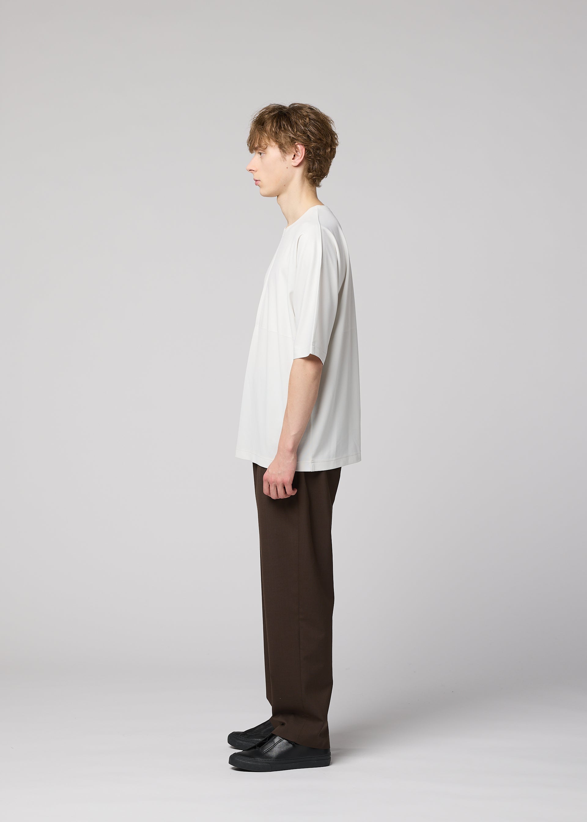 Zoomed view A young person with short light brown hair stands in profile against a plain light background, wearing the IM MEN FLAT T easy-care jersey pressed T-shirt, dark brown pants with bold creases, and black shoes.