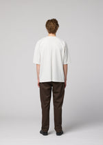 A person with curly hair, seen from behind, wears the IM MEN FLAT T in loose white, dark brown creased pants, and black shoes against a plain light gray background.