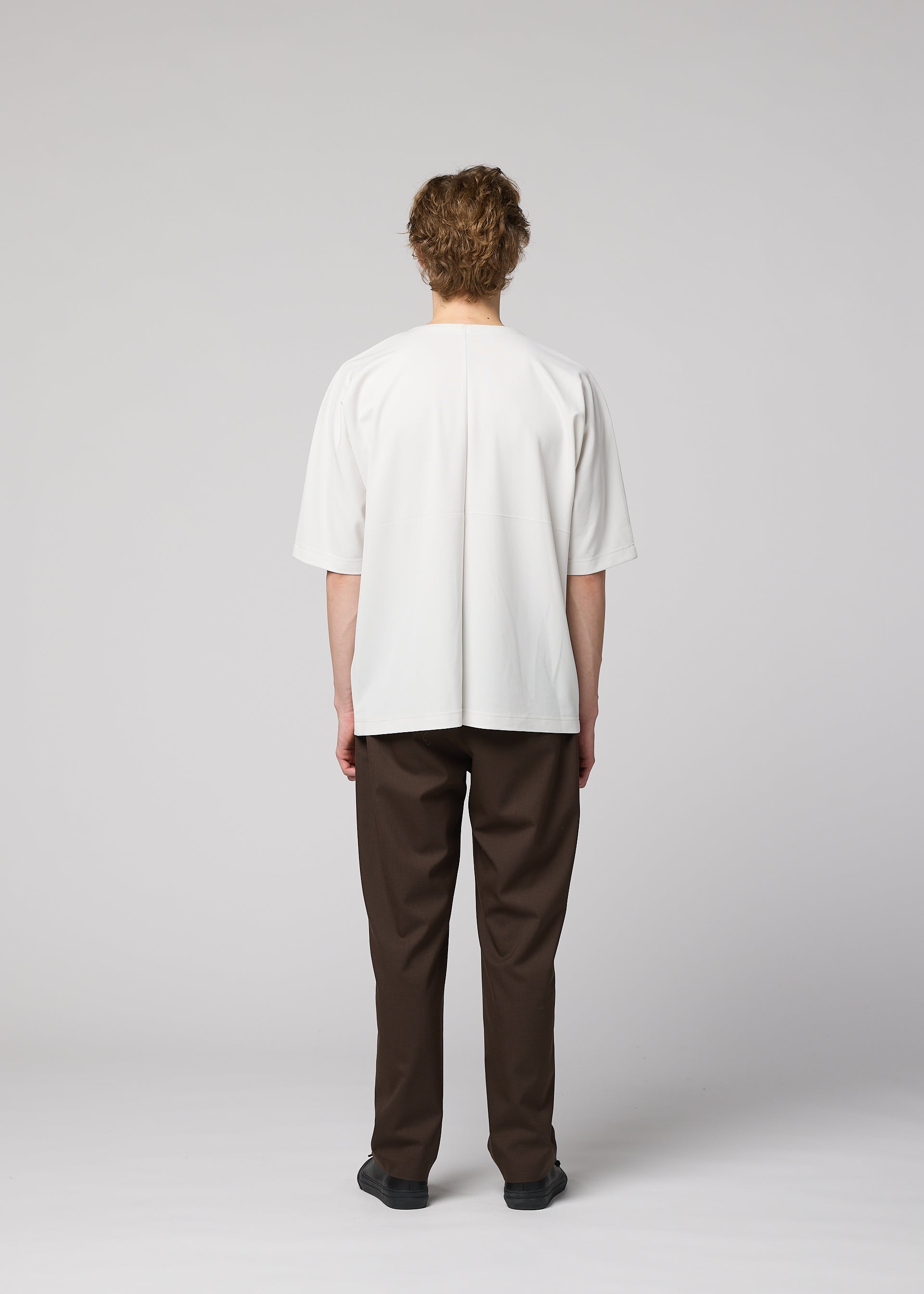 Zoomed view A person with curly hair, seen from behind, wears the IM MEN FLAT T in loose white, dark brown creased pants, and black shoes against a plain light gray background.