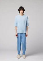 A person with medium-length dark hair models the IM MEN CREPE TUCK JERSEY, a loose, light blue shirt made from wrinkle-resistant jersey fabric, paired with blue pants featuring cinched ankles and white shoes, standing against a plain light gray background.
