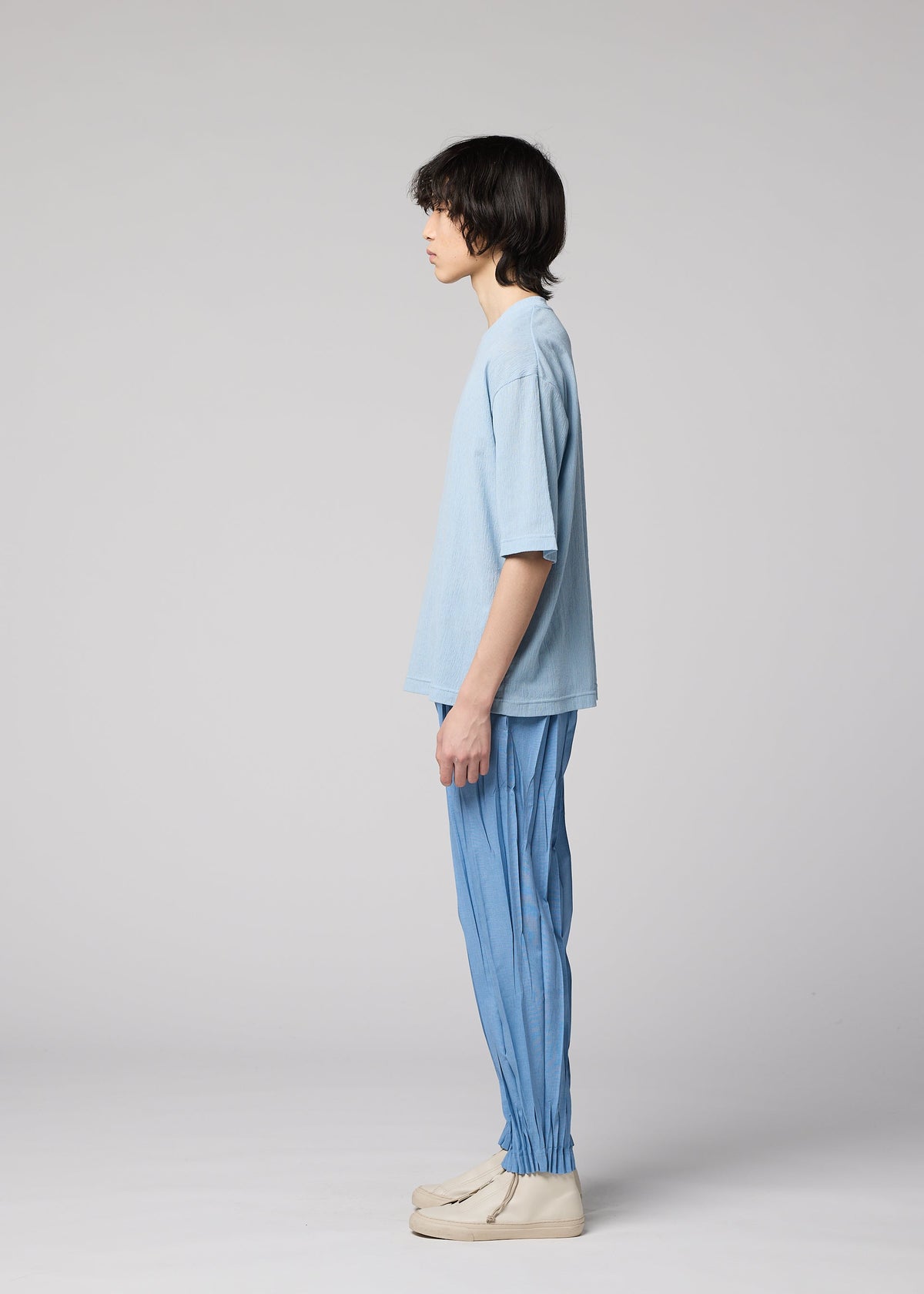 A person with medium-length dark hair stands in profile wearing the IM MEN CREPE TUCK JERSEY, a light blue oversized t-shirt made from wrinkle resistant jersey fabric, paired with blue pleated pants and off-white shoes against a plain light gray background. view 6 of 7