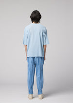 A person with medium-length dark hair, wearing the IM MEN CREPE TUCK JERSEY light blue, quick-drying short-sleeve shirt, loose blue pants with gathered ankles, and light shoes stands facing away from the camera against a plain light background.