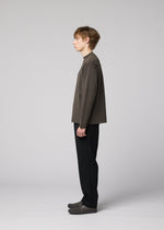 A young person stands in profile against a plain, light background, wearing the IM MEN MONO-MILANO loose, wrinkle-resistant pullover in grey, paired with black pants and grey shoes. They have short, wavy hair and a calm expression.