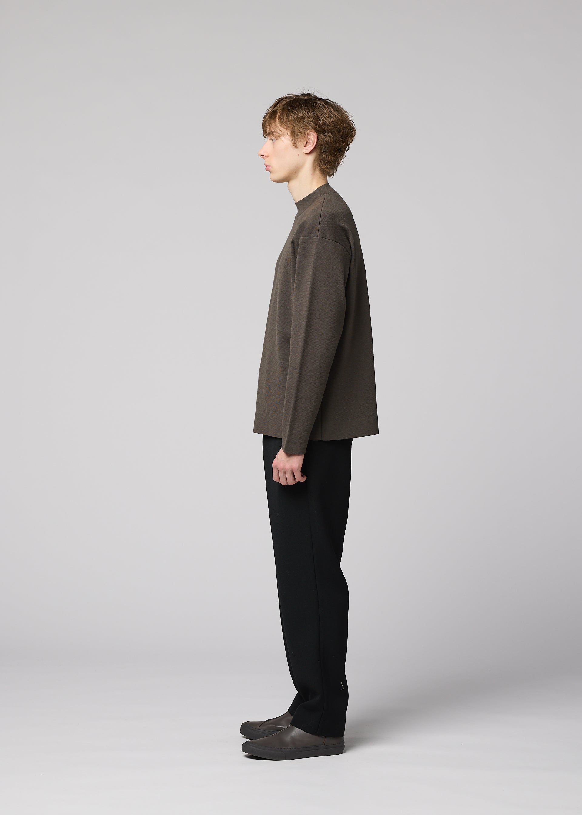 Zoomed view A young person stands in profile against a plain, light background, wearing the IM MEN MONO-MILANO loose, wrinkle-resistant pullover in grey, paired with black pants and grey shoes. They have short, wavy hair and a calm expression.