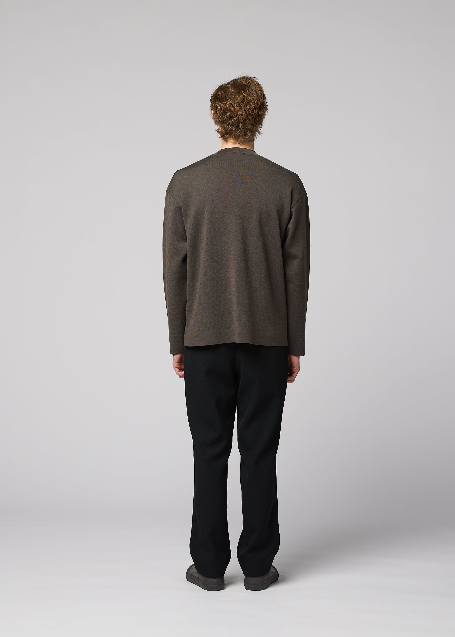 Zoomed view A person stands facing away from the camera, wearing IM MEN’s MONO-MILANO loose dark gray pullover and black pants against a plain light gray background. The wrinkle-resistant fabric of the MONO-MILANO ensures a polished look all day long.