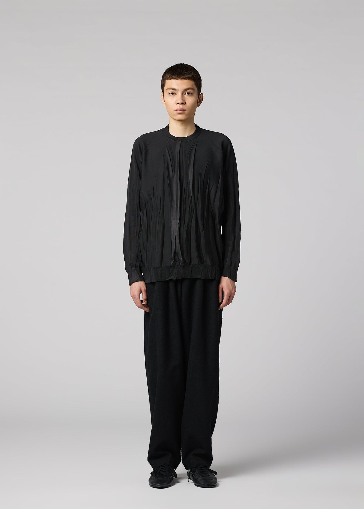 A person stands against a plain light gray background, wearing IM MEN’s RIPPLE KNIT—a black textured polyester long-sleeved top—paired with loose black pants and shoes, looking straight at the camera with a neutral expression. view 4 of 7