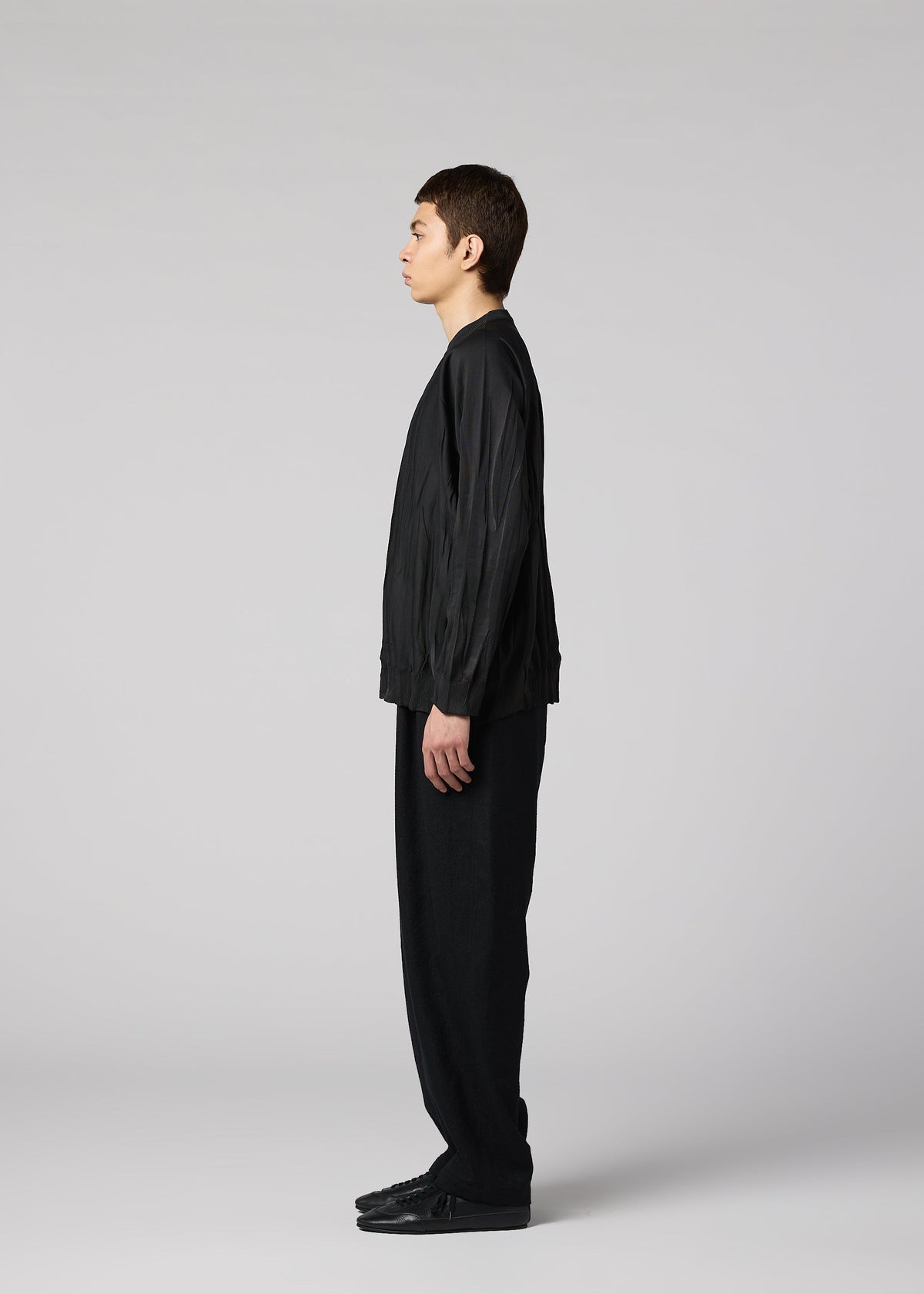 A person with short brown hair stands in profile against a light gray background, wearing IM MEN’s RIPPLE KNIT: a dynamic black long-sleeve top paired with loose black pants and black shoes. view 5 of 7