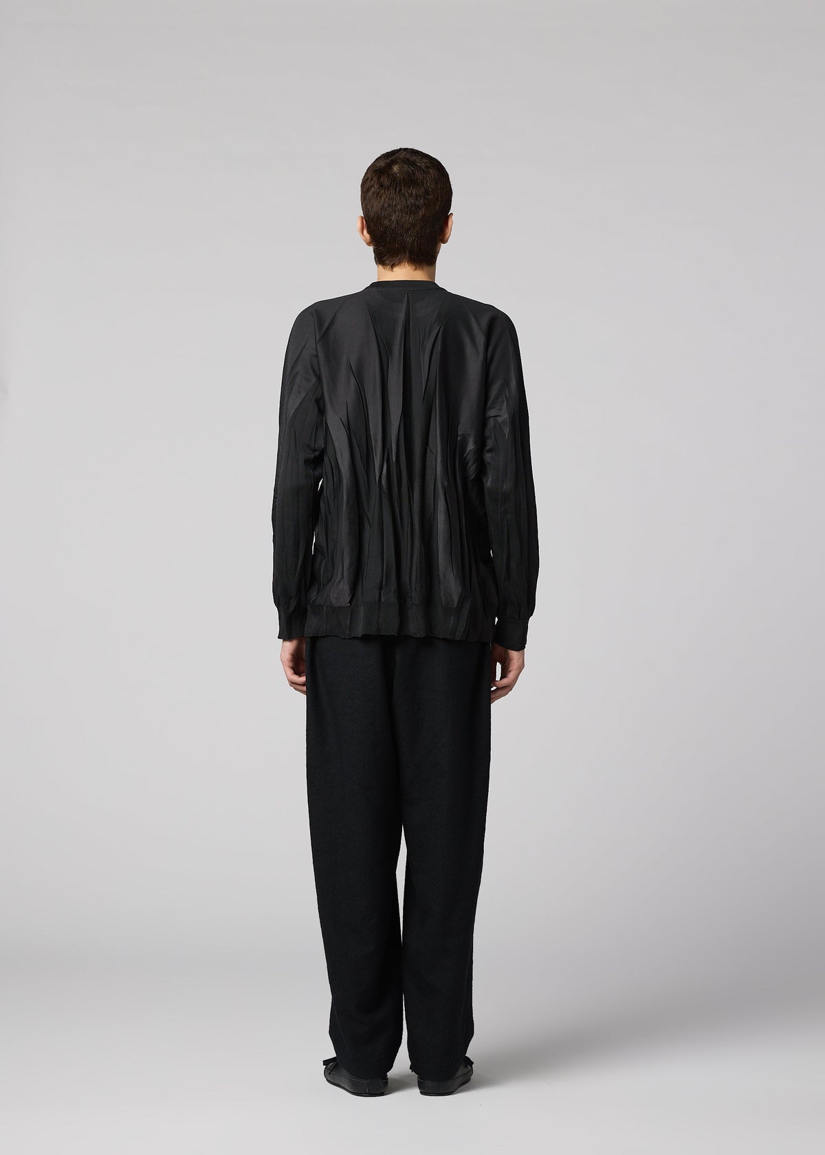 Zoomed view A person stands facing away, wearing the IM MEN RIPPLE KNIT jacket and wide textured polyester pants against a plain light gray background.