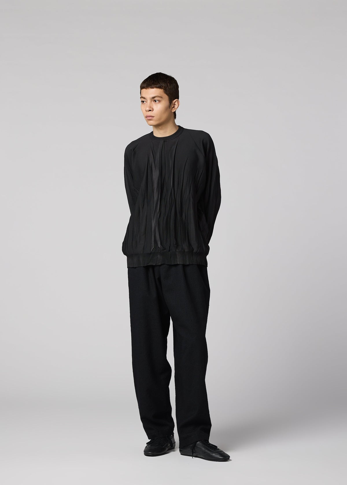 Zoomed view A person stands against a light gray background, hands behind their back, wearing the RIPPLE KNIT by IM MEN—a black pleated textured polyester long-sleeve top—paired with loose black pants and shoes, gazing to the side.
