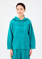 A woman with dark hair tied back smiles gently, wearing the teal SWEATS PLEATS HOOD by me ISSEY MIYAKE—a pleated, jersey fleece-lined hoodie—while standing against a plain white background.