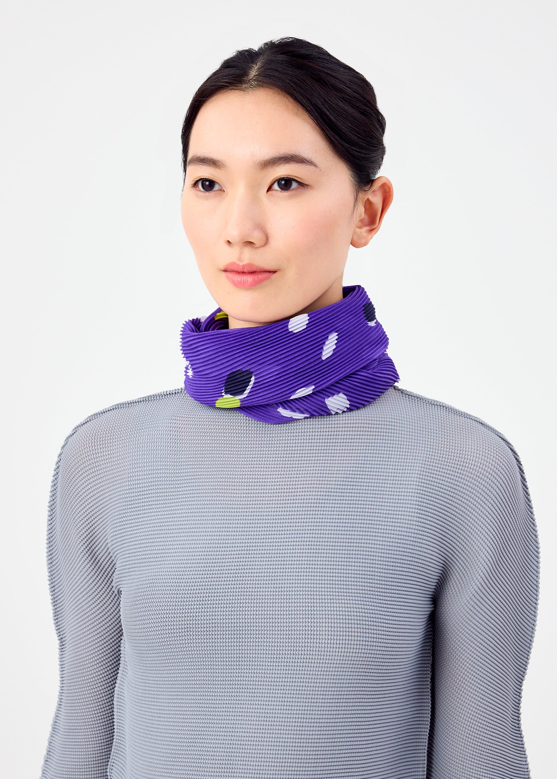 Zoomed view A woman with dark hair tied back wears a light gray textured top and the TIME DOTS PLEATS STOLE by me ISSEY MIYAKE, featuring purple pleats with white and black patterns, wrapped around her neck against a plain white background.