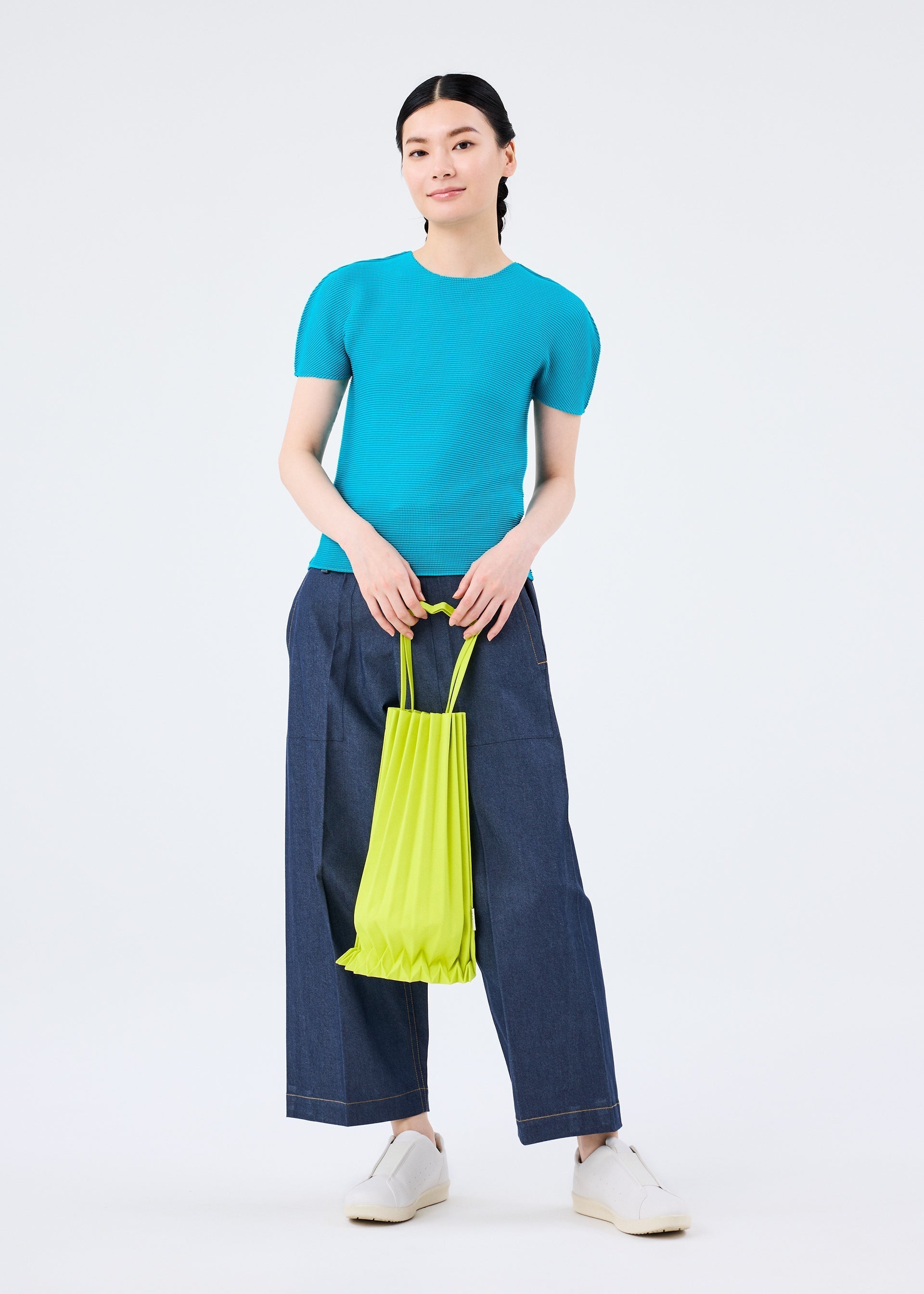 Zoomed view A person stands against a white background, wearing a turquoise top, wide-leg dark denim pants, white shoes, and holding a bright yellow TRUNK PLEATS BAG 12 by me ISSEY MIYAKE, known for its unique handle and vivid color variations.