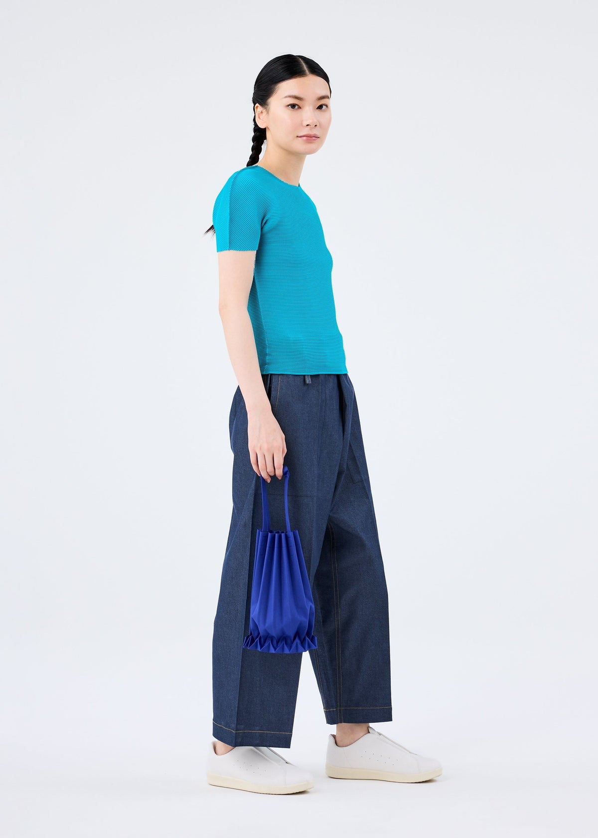 A person stands against a plain white background, wearing a turquoise short-sleeve top, wide-leg blue jeans, white sneakers, and holding the vivid TRUNK PLEATS BAG 12 by me ISSEY MIYAKE at their side. view 14 of 14