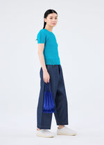 A person stands against a plain white background, wearing a turquoise short-sleeve top, wide-leg blue jeans, white sneakers, and holding the vivid TRUNK PLEATS BAG 12 by me ISSEY MIYAKE at their side.