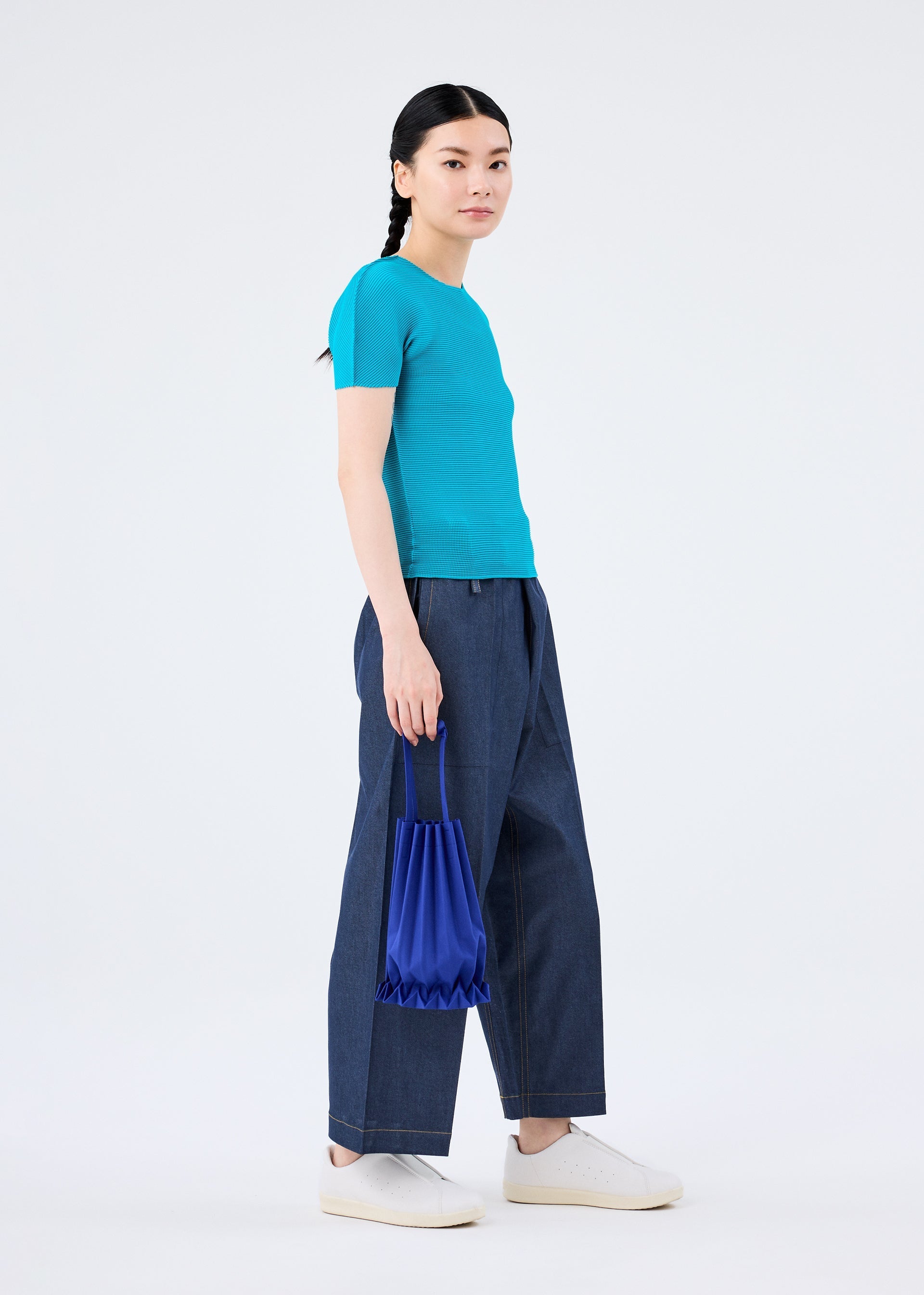 Zoomed view A person stands against a plain white background, wearing a turquoise short-sleeve top, wide-leg blue jeans, white sneakers, and holding the vivid TRUNK PLEATS BAG 12 by me ISSEY MIYAKE at their side.