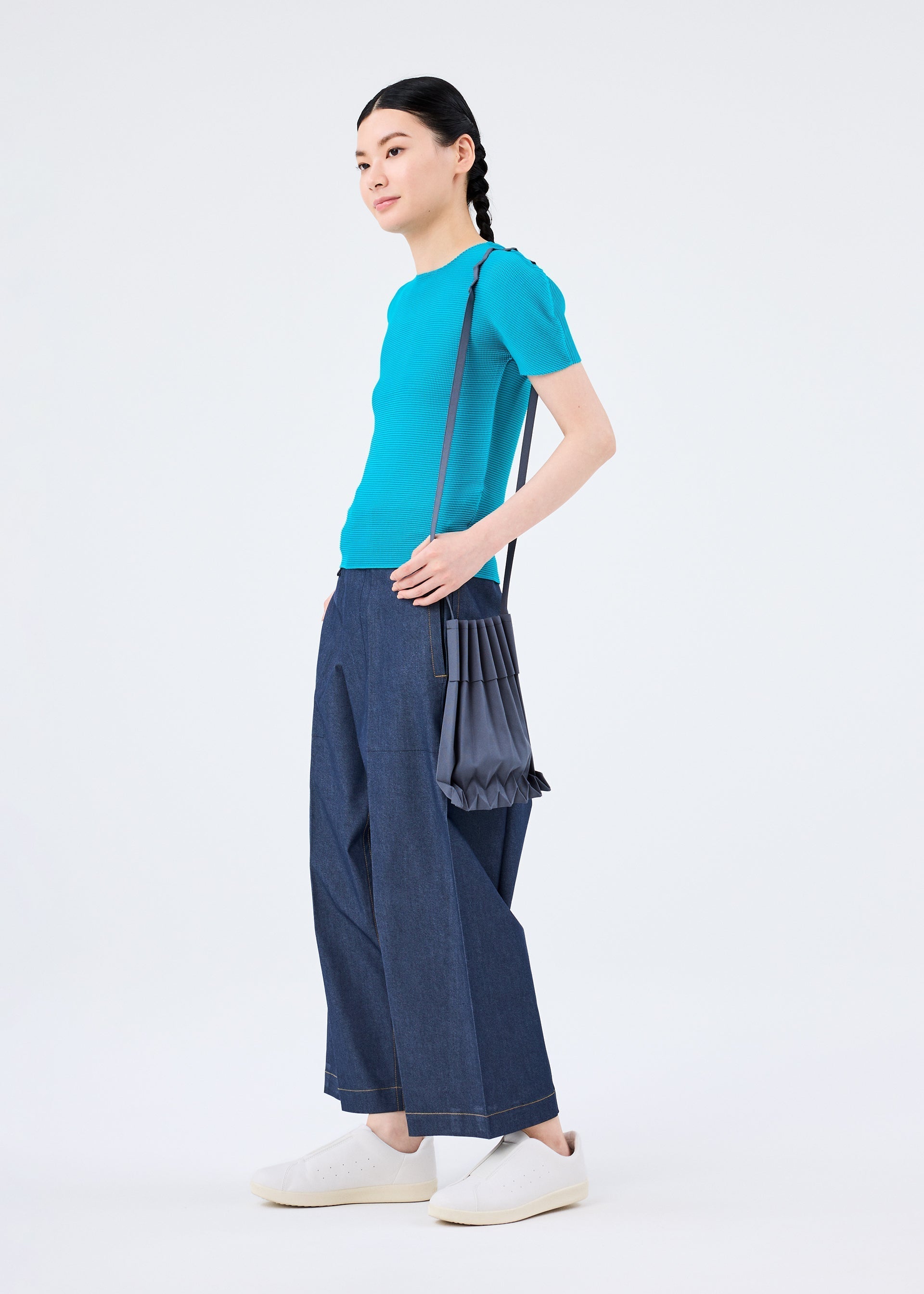 Zoomed view A person with braided hair stands against a plain white background, wearing a bright blue short-sleeve top, wide-leg dark blue jeans, white sneakers, and carrying the me ISSEY MIYAKE TRUNK PLEATS BAG 12 over their shoulder.