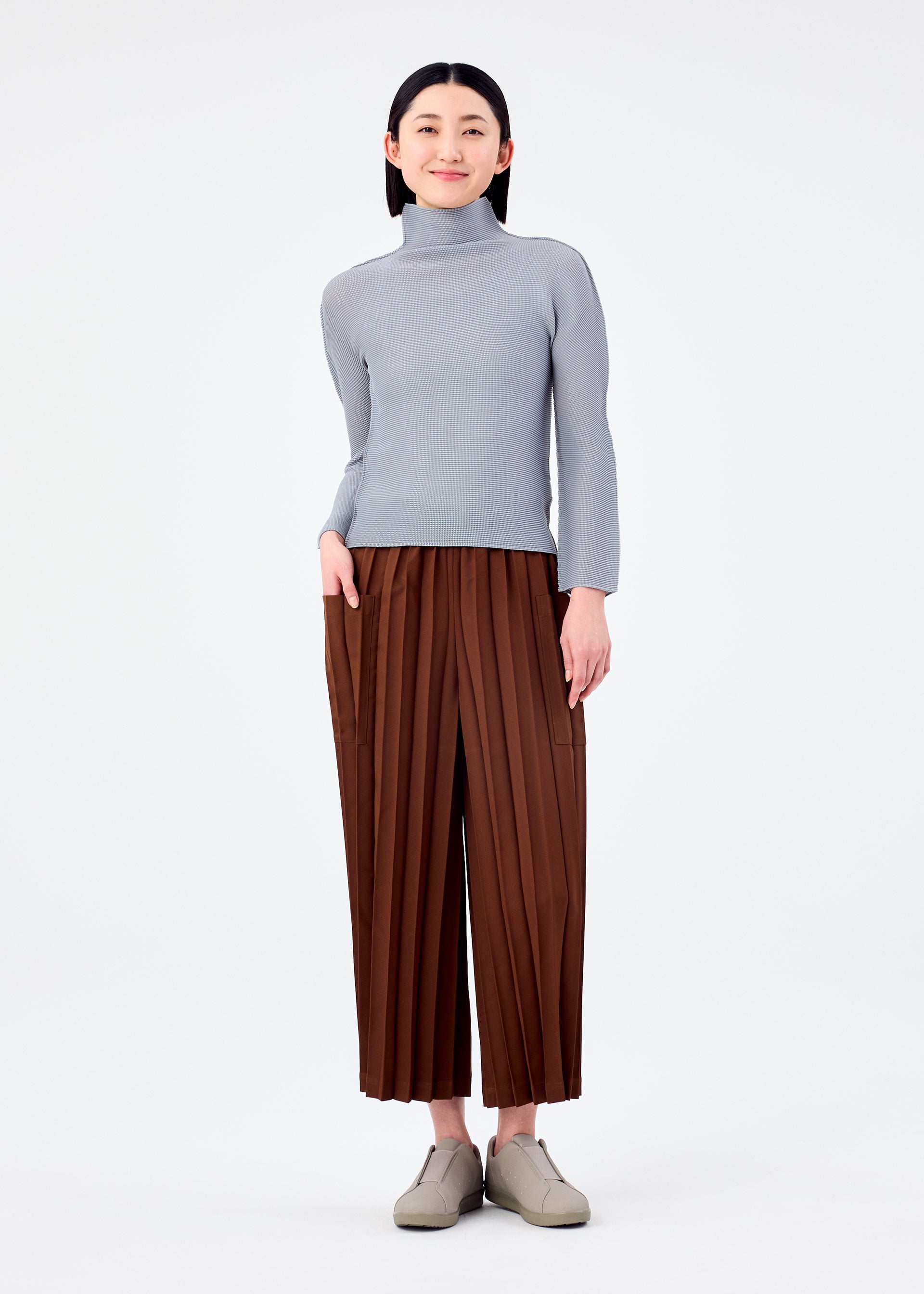 Zoomed view A person with straight dark hair smiles gently, wearing me ISSEY MIYAKE's COLOR PLEATS BOTTOM wide brown pleated pants with pockets, a light gray turtleneck, and beige shoes against a plain white background.