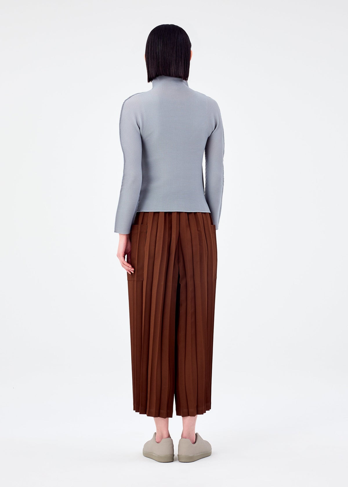 A person stands facing away, wearing a light gray long-sleeve top, COLOR PLEATS BOTTOM brown pleated pants with pockets by me ISSEY MIYAKE, and light slip-on shoes against a plain white background. view 7 of 7