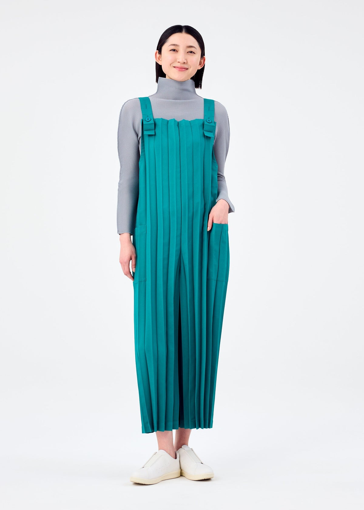 A person stands against a white background wearing the me ISSEY MIYAKE COLOR PLEATS BOTTOM—a teal pleated jumpsuit with pockets—paired with a gray turtleneck top and white slip-on shoes, one hand in their pocket and a neutral expression. view 6 of 7