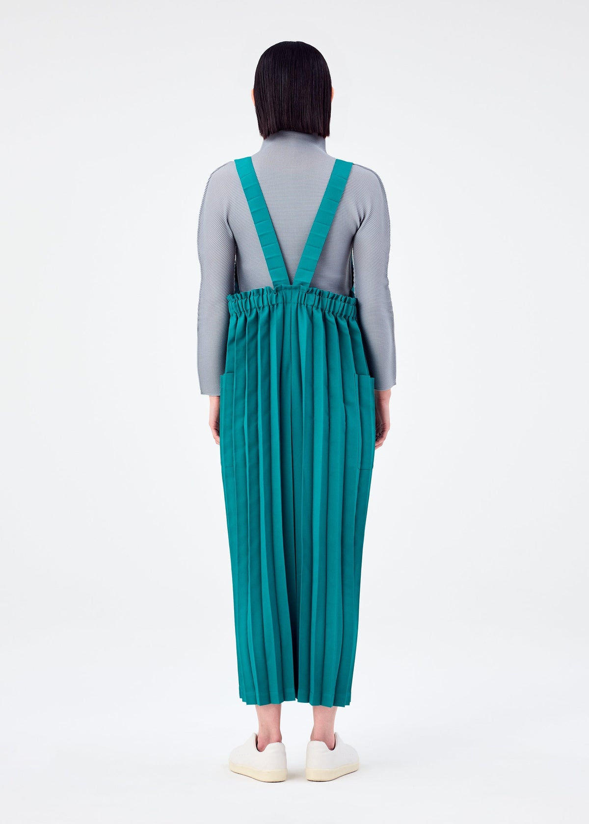 A person faces away, dressed in a grey long-sleeve top and me ISSEY MIYAKE COLOR PLEATS BOTTOM—a green pleated jumpsuit with an elastic waist and suspender style—paired with white shoes against a plain white background. view 7 of 7