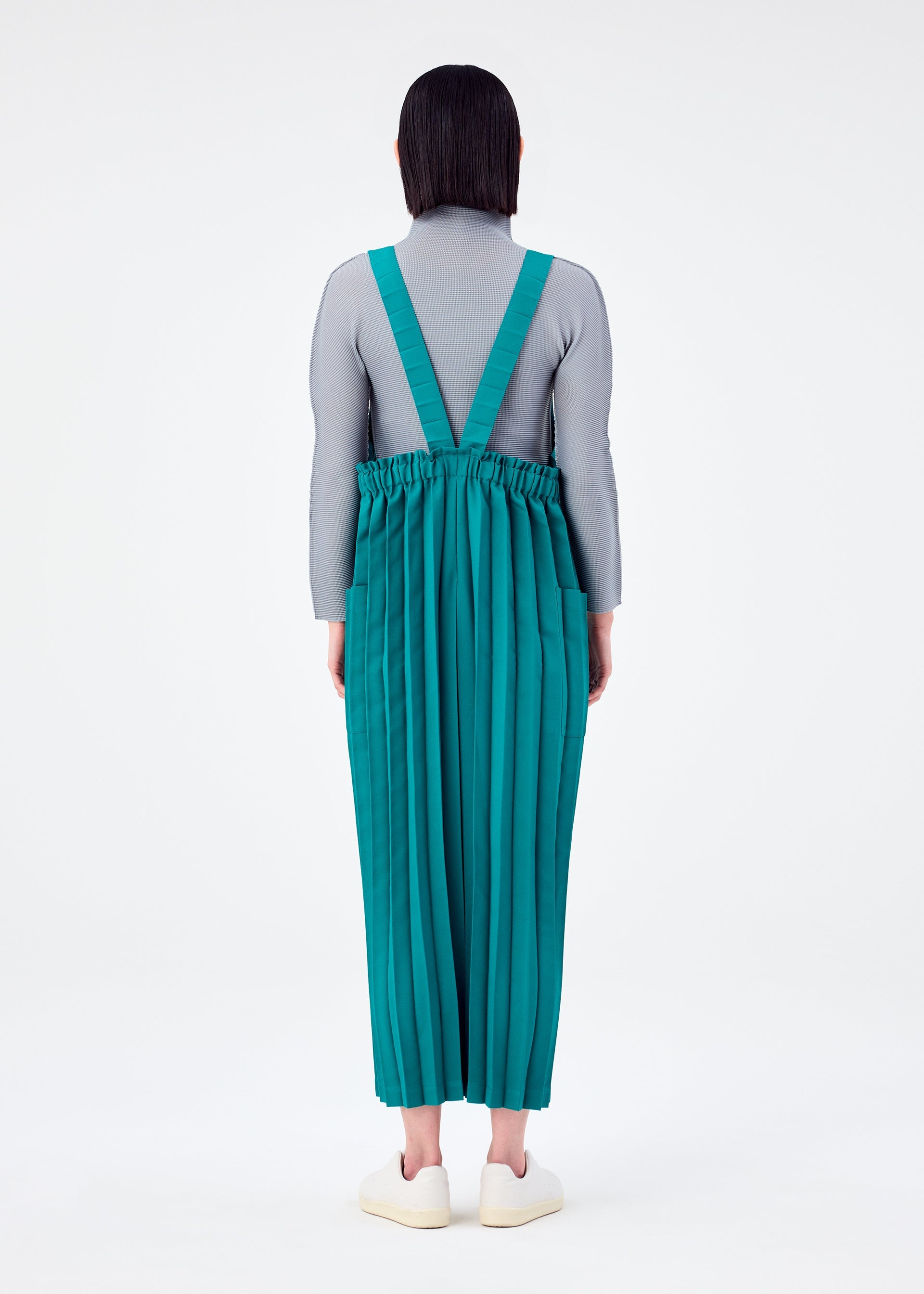 Zoomed view A person faces away, dressed in a grey long-sleeve top and me ISSEY MIYAKE COLOR PLEATS BOTTOM—a green pleated jumpsuit with an elastic waist and suspender style—paired with white shoes against a plain white background.