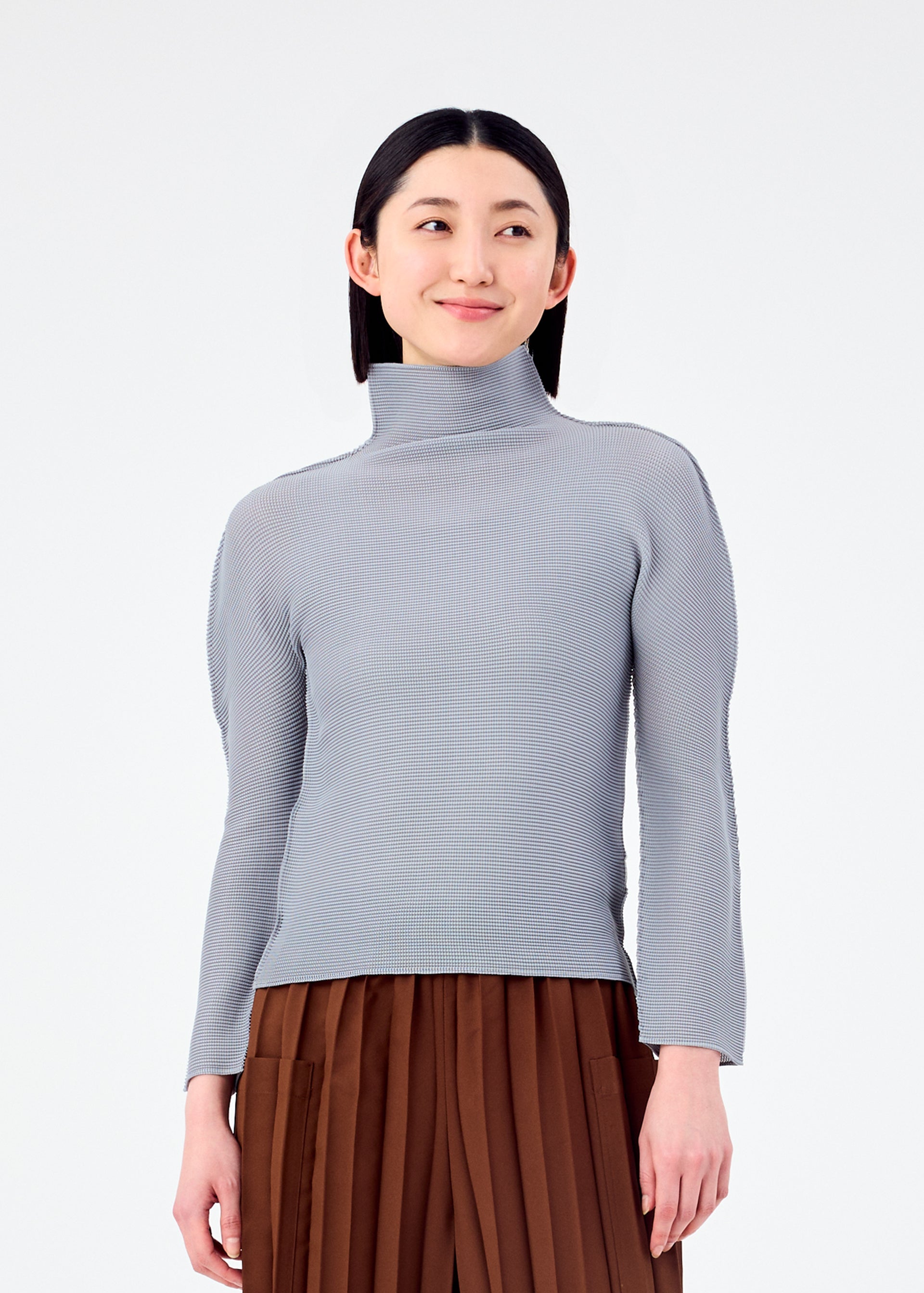 Zoomed view A woman with straight dark hair, dressed in the me ISSEY MIYAKE STRETCH PLEATS 7 light gray pleated long-sleeve top and brown pleated pants, stands against a white background and smiles slightly while looking to her left.