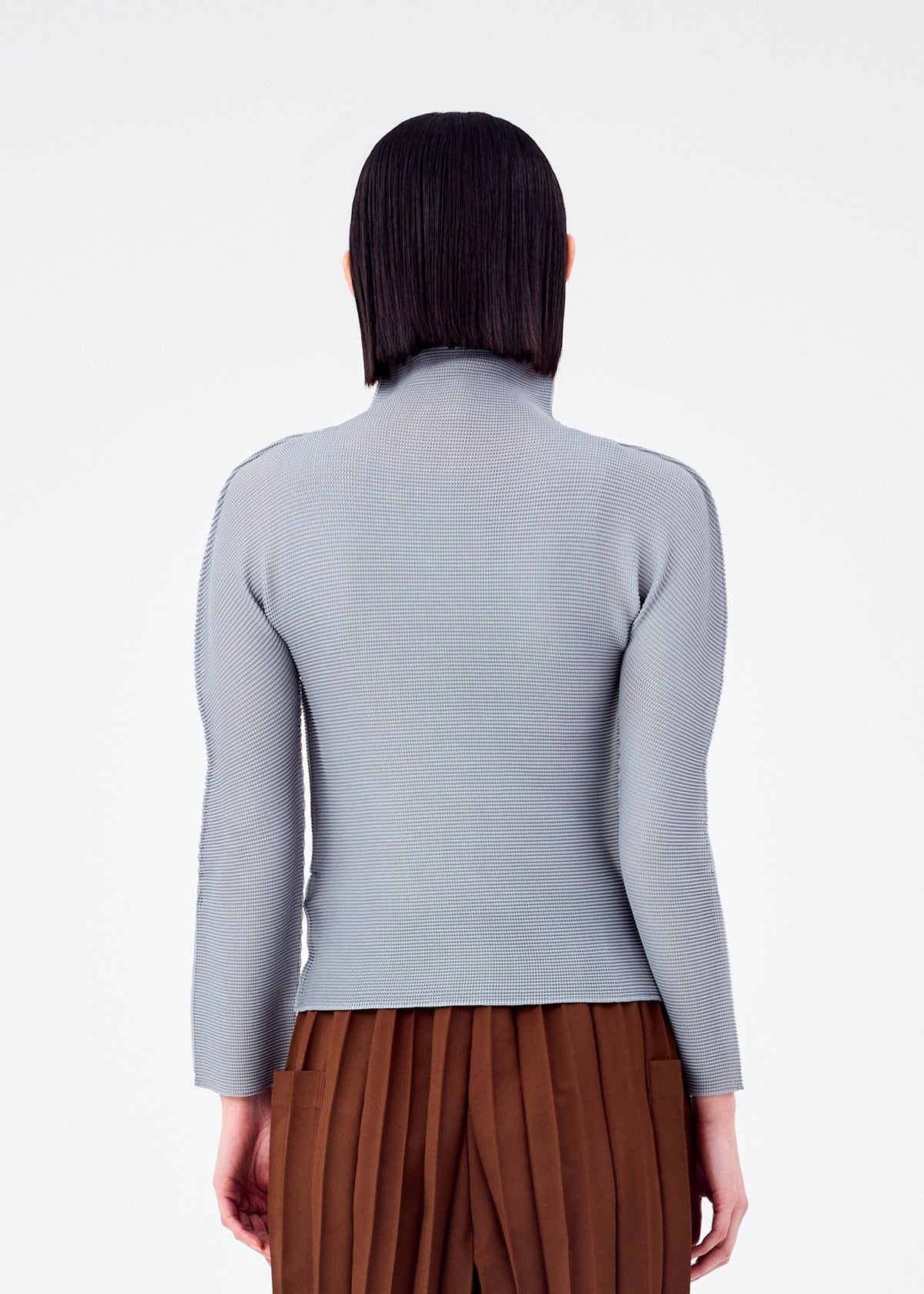 A person with straight, dark hair faces away from the camera, wearing a light gray STRETCH PLEATS 7 long-sleeve top and brown pleated pants by me ISSEY MIYAKE, set against a plain white background. view 6 of 7