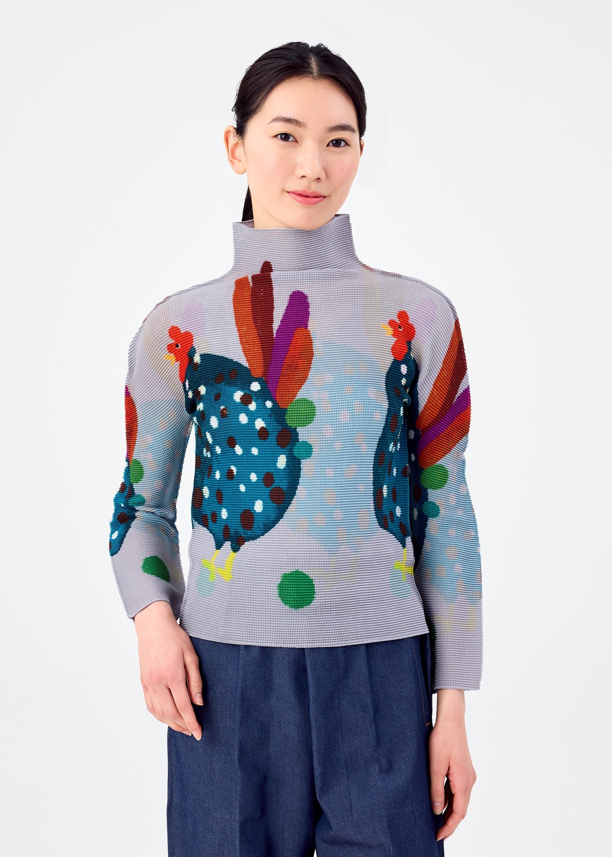 A woman with straight dark hair wears the DAWN high-neck top by me ISSEY MIYAKE, featuring colorful abstract chicken designs and paired with loose blue pants, standing against a white background and smiling softly. view 4 of 6