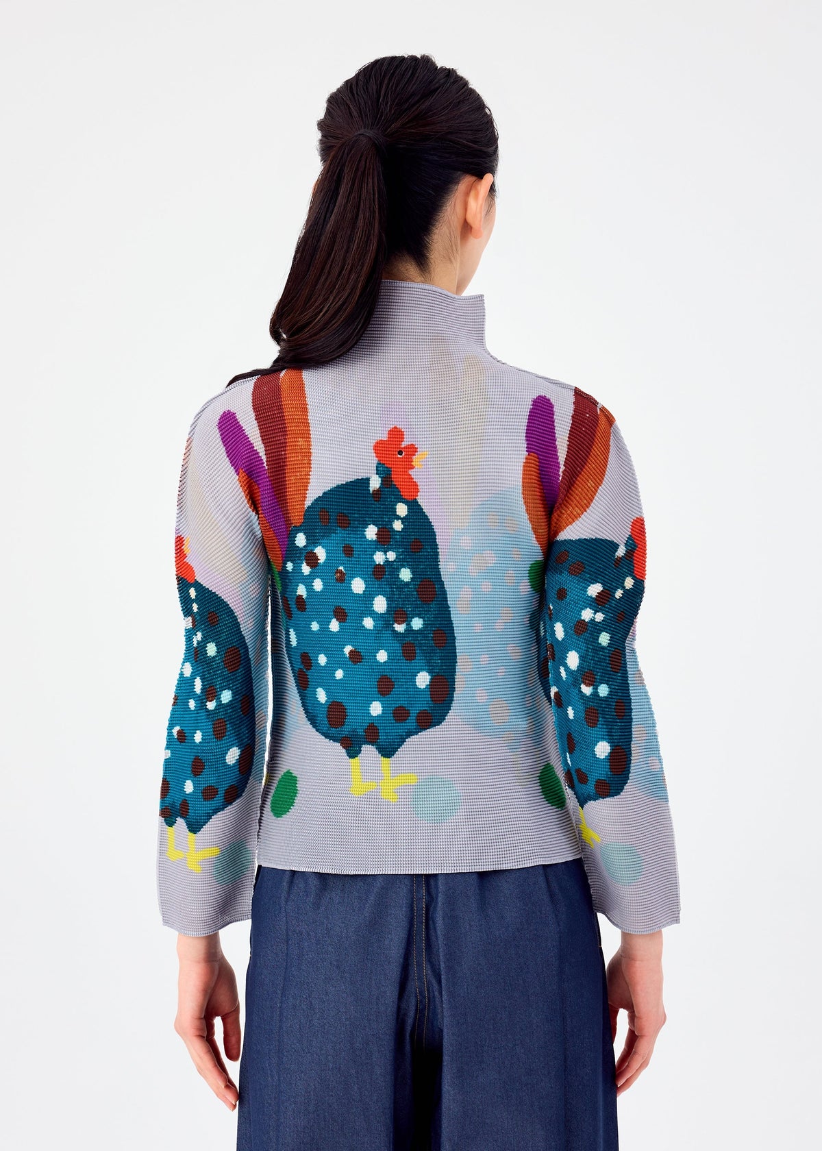 A woman with dark hair in a ponytail is seen from behind, wearing DAWN by me ISSEY MIYAKE—an elegant light gray pleated top with bold blue, orange, and purple chicken designs—paired with blue wide-leg pants. view 5 of 6