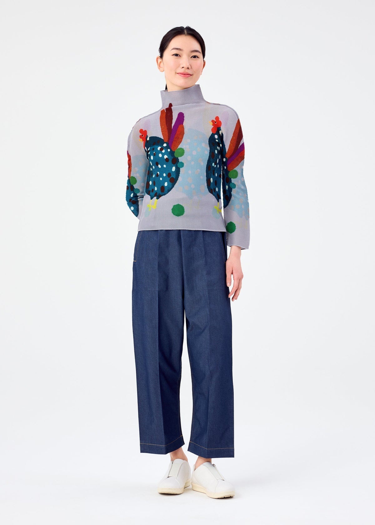 A woman stands against a white background, smiling softly with her hands at her sides, wearing the DAWN high-neck top by me ISSEY MIYAKE featuring colorful chicken designs, paired with loose blue trousers and white slip-on shoes. view 6 of 6