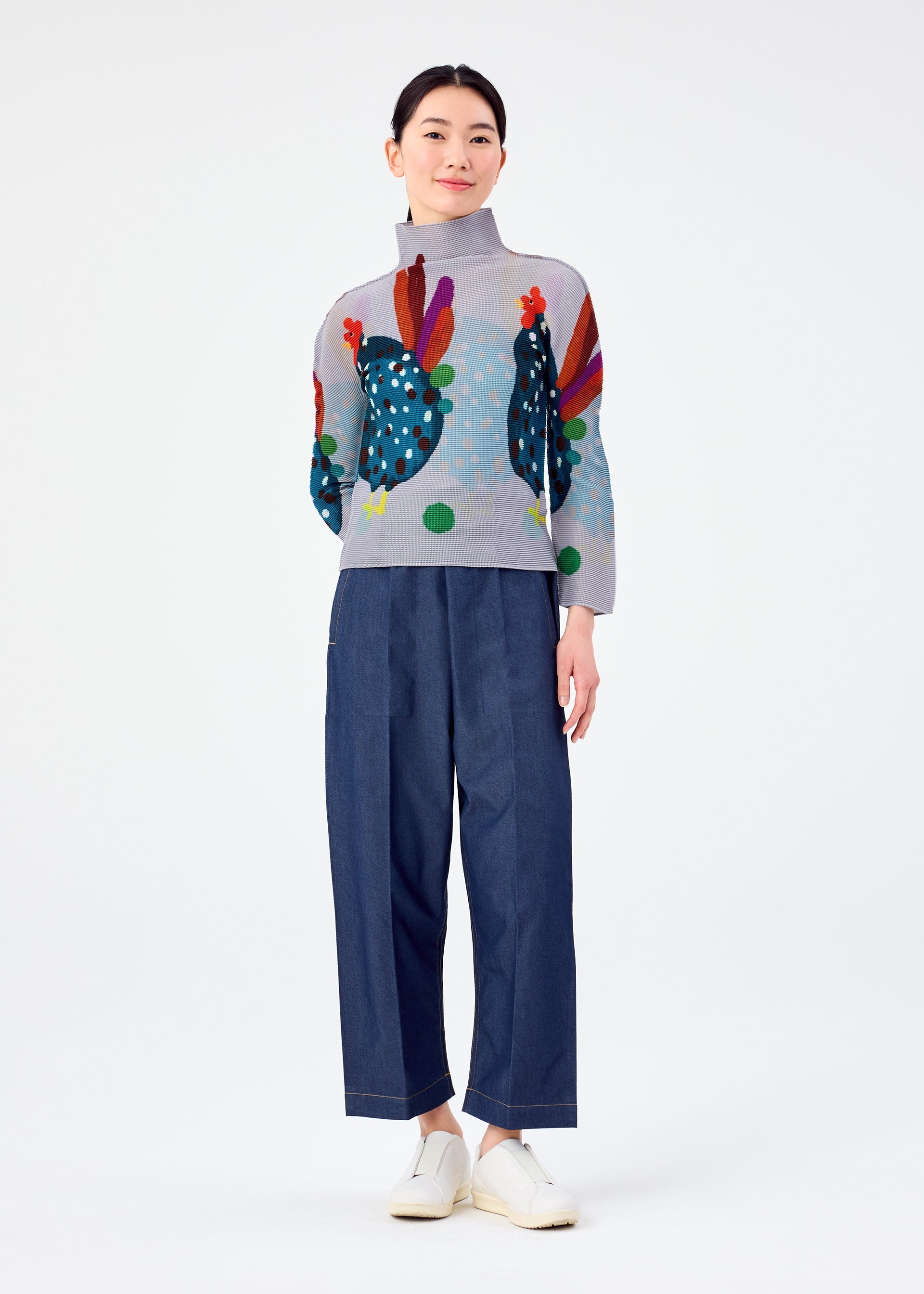 Zoomed view A woman stands against a white background, smiling softly with her hands at her sides, wearing the DAWN high-neck top by me ISSEY MIYAKE featuring colorful chicken designs, paired with loose blue trousers and white slip-on shoes.