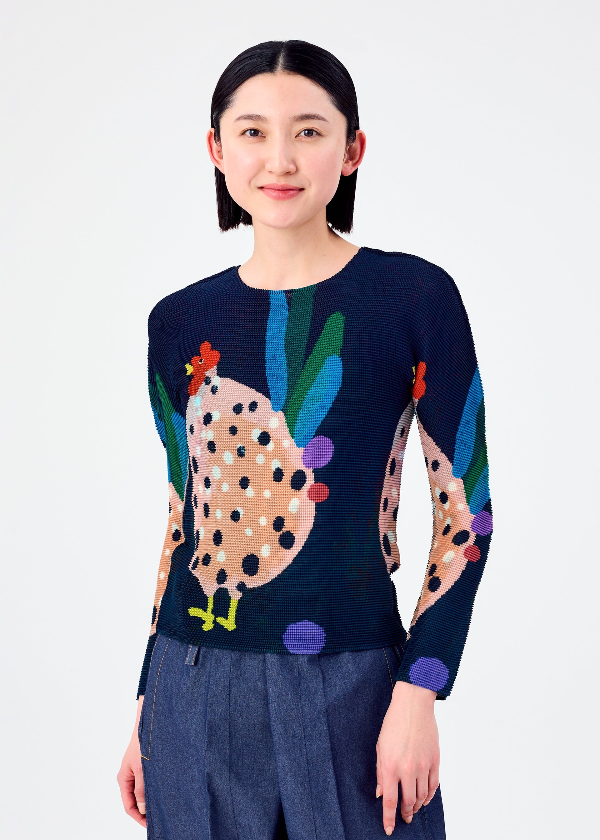 Zoomed view A woman with straight dark hair smiles in the DAWN top by me ISSEY MIYAKE—a navy, long-sleeved stretchy shirt with a colorful abstract chicken pattern—paired with wide-leg blue pants against a plain white background.