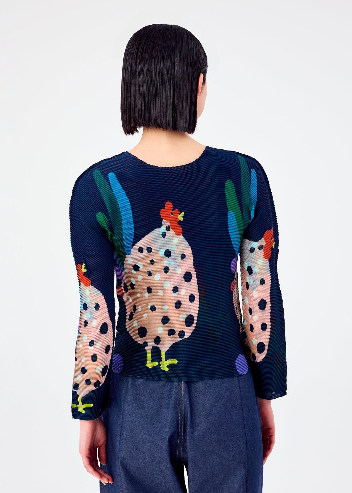 A person with straight, dark, chin-length hair is seen from behind, wearing the me ISSEY MIYAKE DAWN long-sleeved stretchy top with a colorful chicken and polka dot pattern, paired with dark blue pants against a plain white background. view 5 of 6