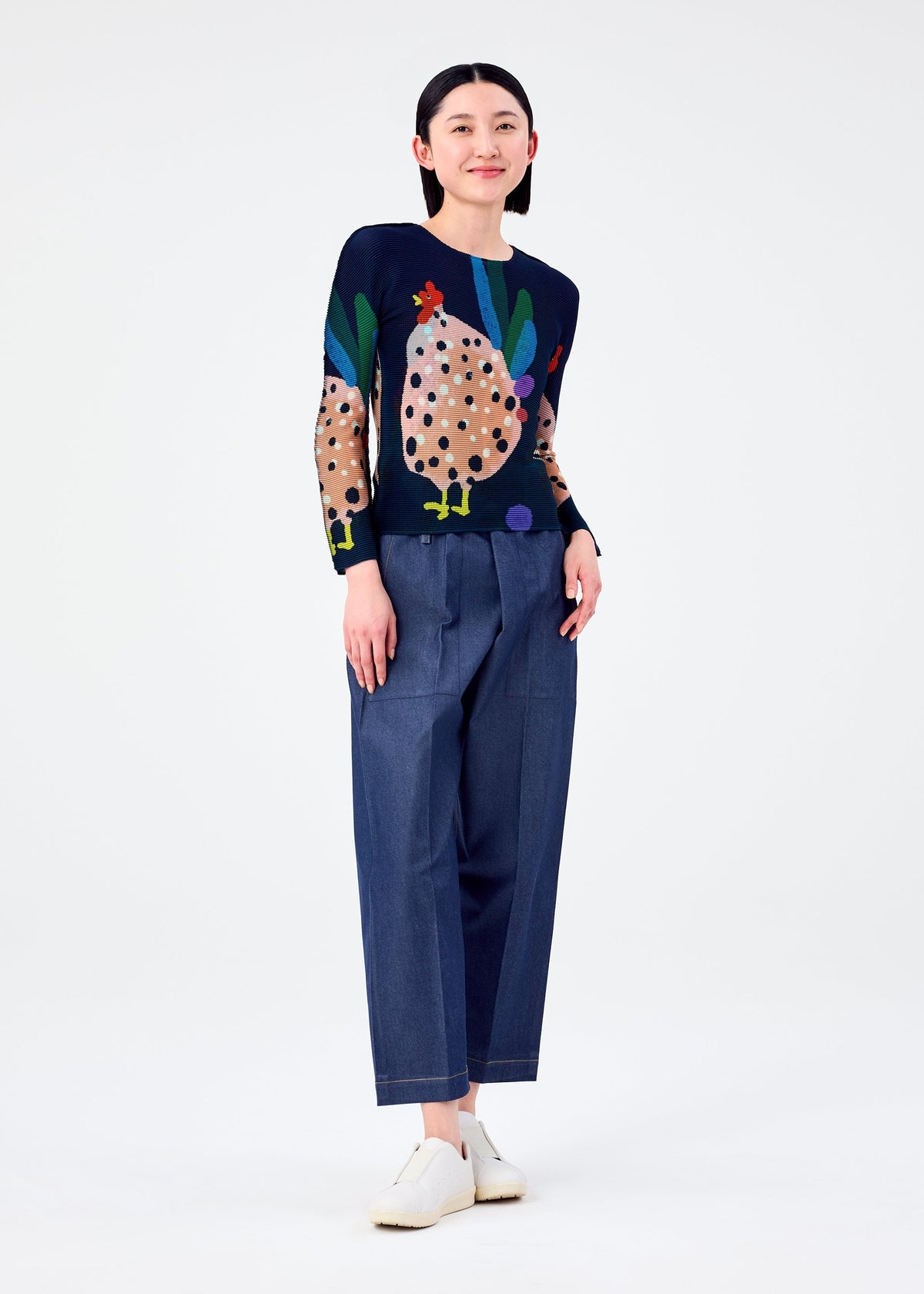 A smiling person with straight black hair wears DAWN, a long-sleeved stretchy top with a colorful chicken print by me ISSEY MIYAKE, paired with wide-leg blue pants and white slip-on shoes against a plain white background. view 6 of 6