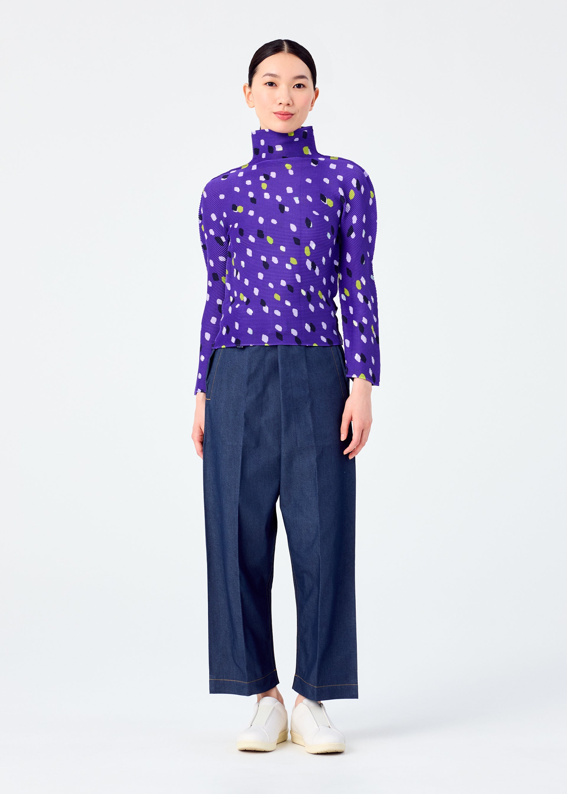 Zoomed view A person faces forward in the me ISSEY MIYAKE TIME DOTS high-neck, long-sleeved top featuring a purple, white, and green dotted pattern, paired with wide-leg dark blue pants and white shoes against a plain white background.