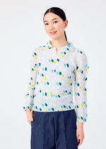 A woman with straight dark hair in a ponytail wears the TIME DOTS long-sleeved pleated top by me ISSEY MIYAKE, featuring a blue and yellow dotted pattern, paired with dark blue pants. She stands smiling against a plain white background with hands in her pockets.