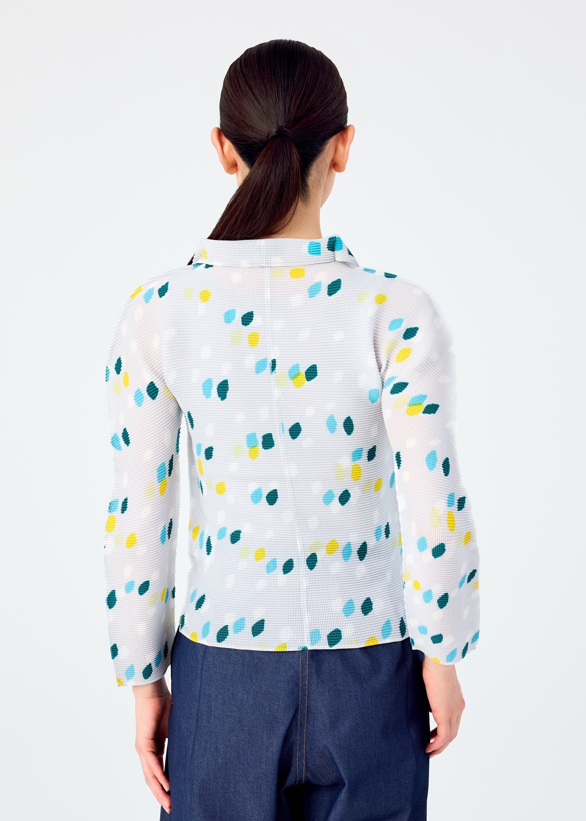 Zoomed view A person with long dark hair in a low ponytail is shown from behind, wearing the TIME DOTS white long-sleeved pleated top with blue, green, and yellow dot patterns by me ISSEY MIYAKE, paired with dark blue pants against a plain white background.