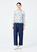 A person faces forward against a plain white background, wearing TIME DOTS by me ISSEY MIYAKE—a long-sleeved pleated top with colorful dots—paired with loose dark blue pants and white shoes.