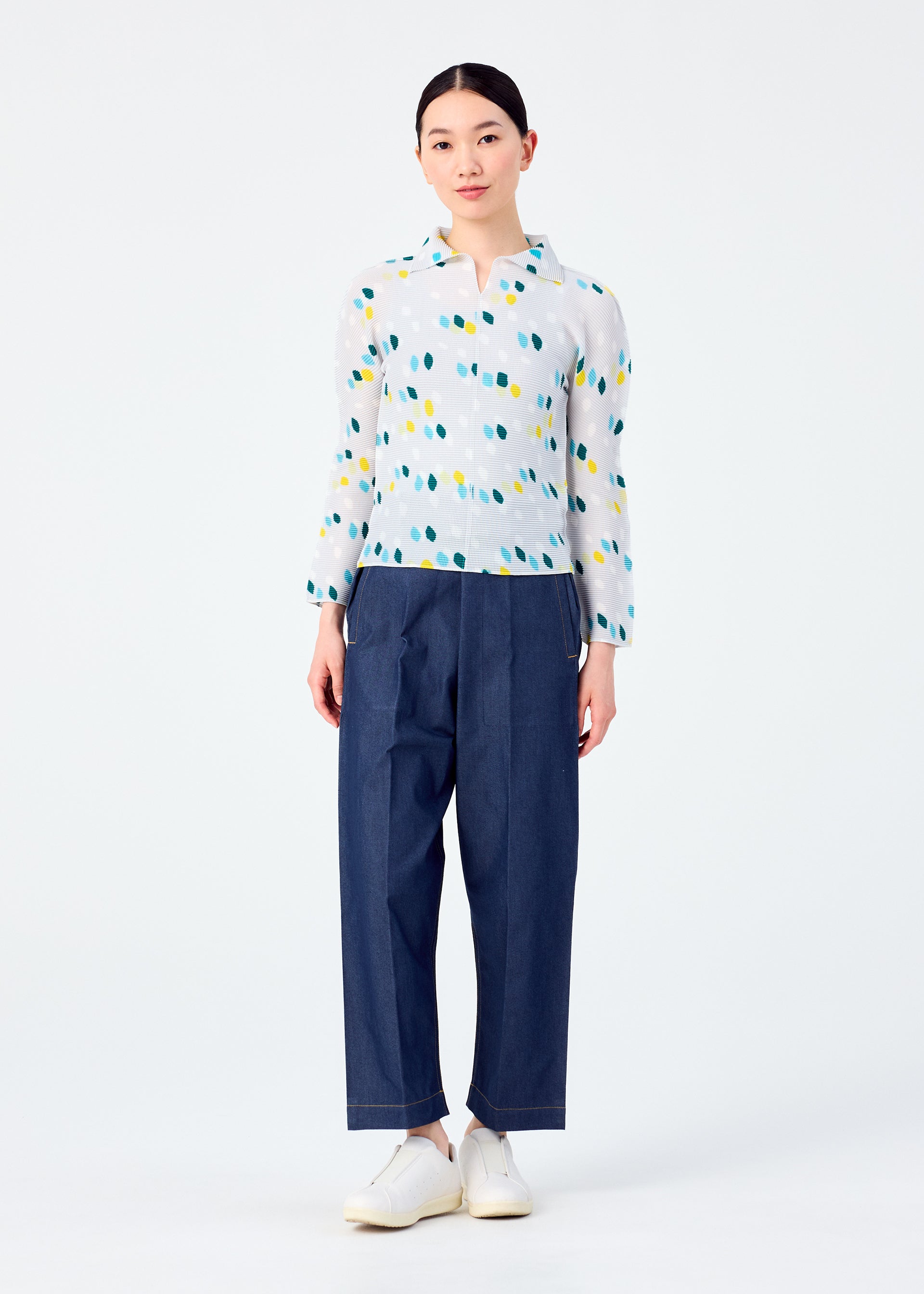 Zoomed view A person faces forward against a plain white background, wearing TIME DOTS by me ISSEY MIYAKE—a long-sleeved pleated top with colorful dots—paired with loose dark blue pants and white shoes.