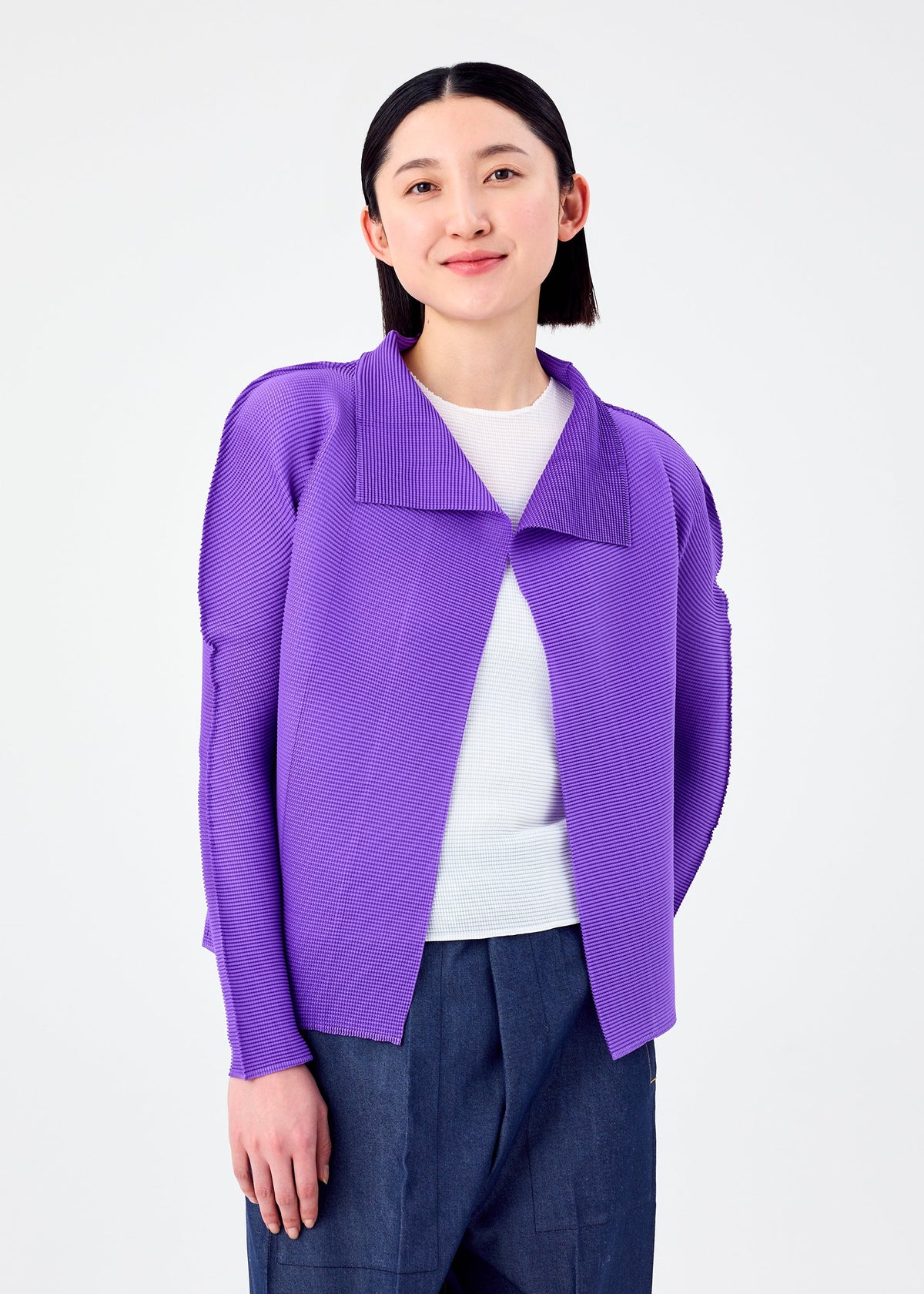 A woman with straight black hair, dressed in a bright purple me ISSEY MIYAKE STRETCH PLEATS 7 cardigan over a white top and blue pants, stands against a plain white background and smiles softly at the camera. view 5 of 7