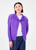 A woman with straight black hair, dressed in a bright purple me ISSEY MIYAKE STRETCH PLEATS 7 cardigan over a white top and blue pants, stands against a plain white background and smiles softly at the camera.