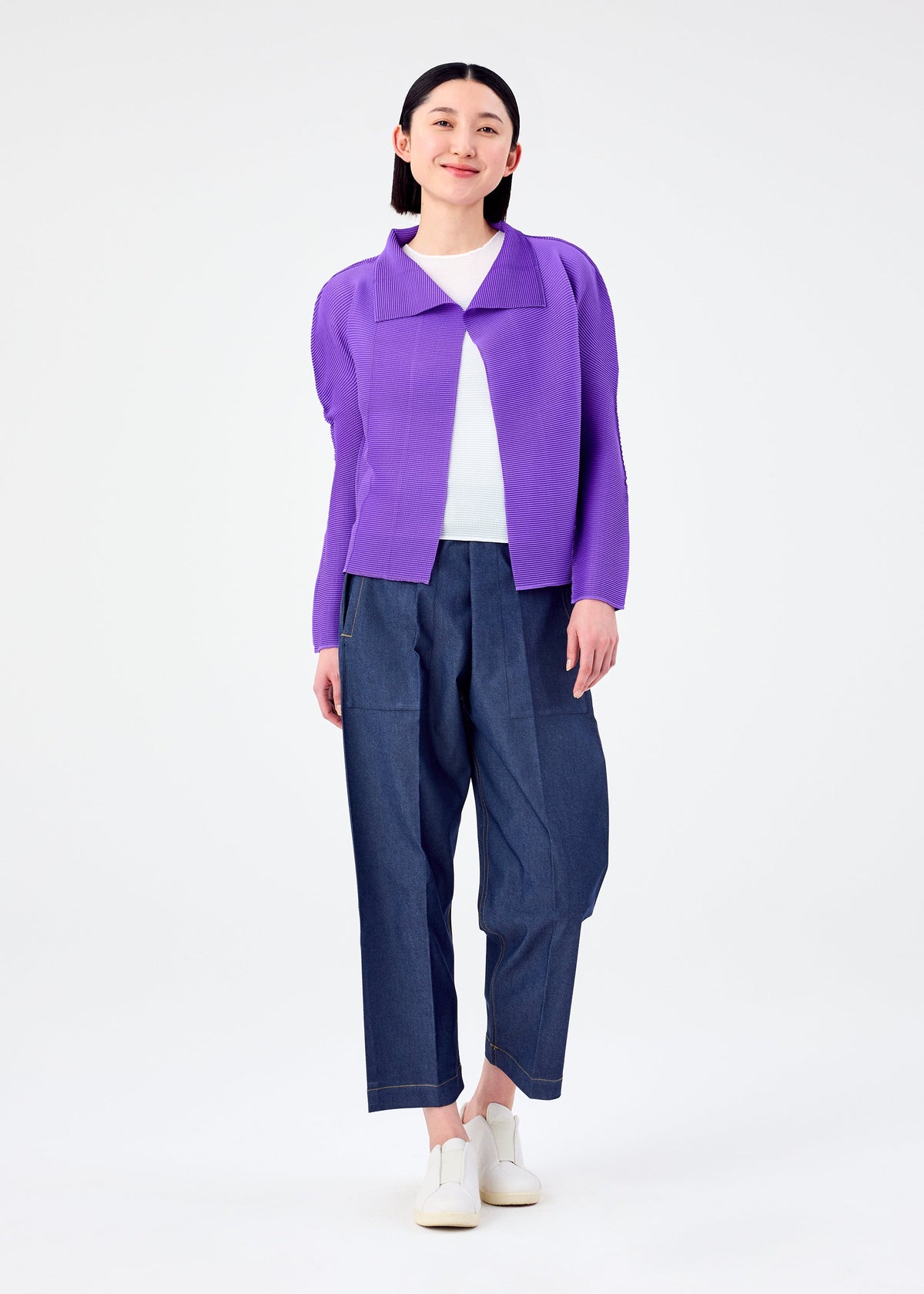 A person smiles against a plain white background, wearing a purple me ISSEY MIYAKE STRETCH PLEATS 7 long-sleeved cardigan over a white top, loose blue pants, and white shoes. view 7 of 7
