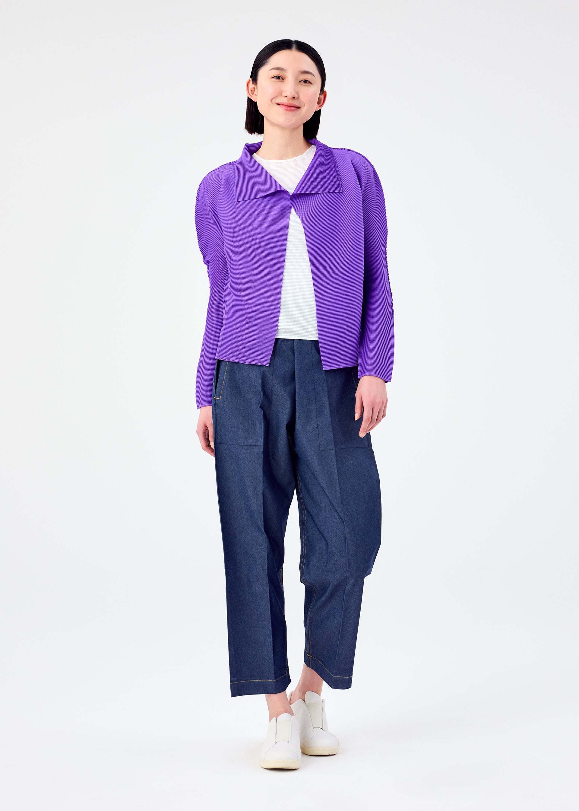 Zoomed view A person smiles against a plain white background, wearing a purple me ISSEY MIYAKE STRETCH PLEATS 7 long-sleeved cardigan over a white top, loose blue pants, and white shoes.