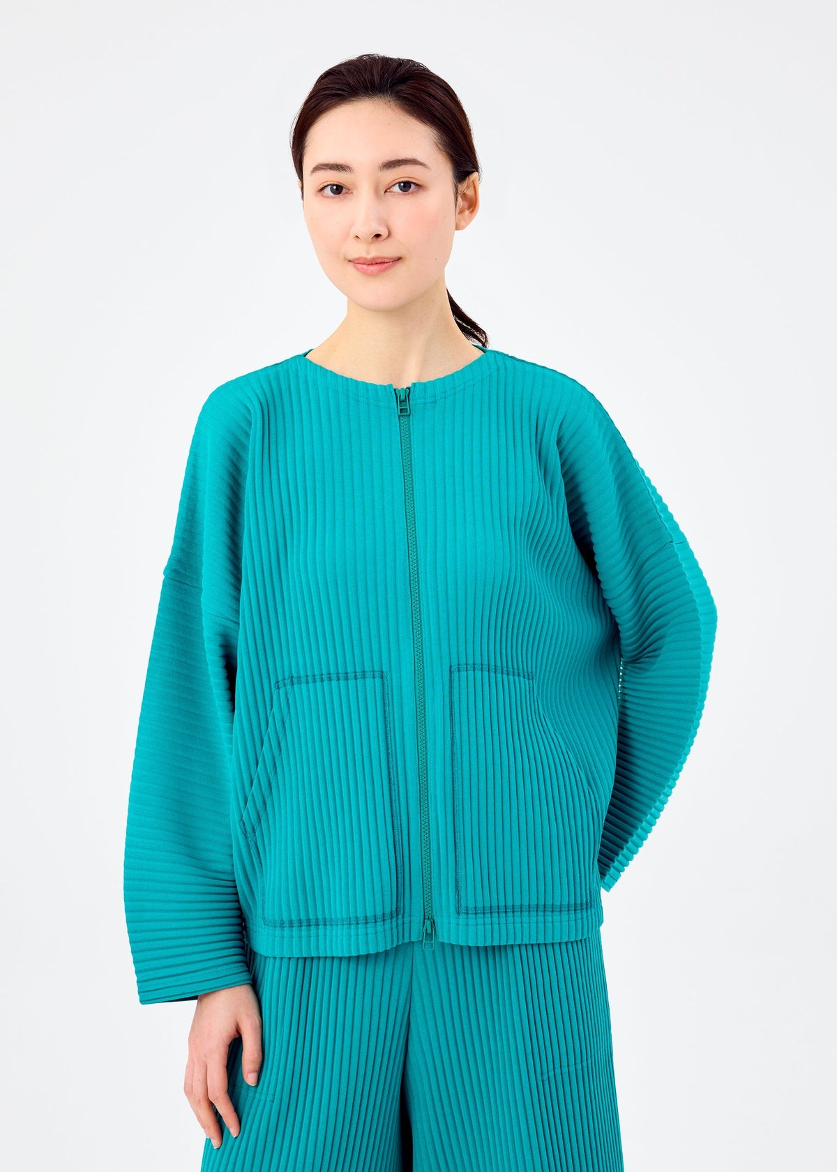 A woman with long dark hair pulled back wears the me ISSEY MIYAKE SWEATS PLEATS—a teal, pleated cardigan with matching pants—standing against a plain white background and facing forward with a slight smile. view 5 of 7