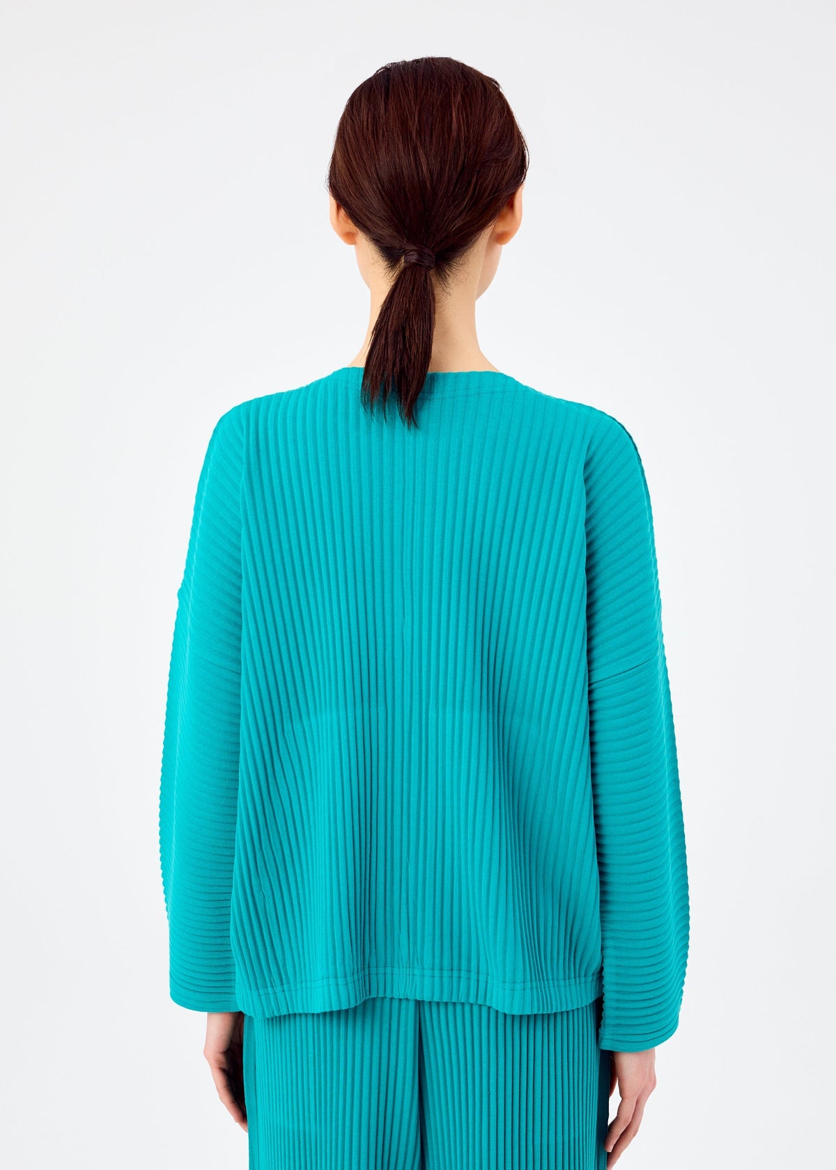A person with dark brown hair tied back is seen from behind, wearing the bright teal SWEATS PLEATS long-sleeved pleated cardigan and matching pleated pants by me ISSEY MIYAKE, against a plain white background. view 6 of 7